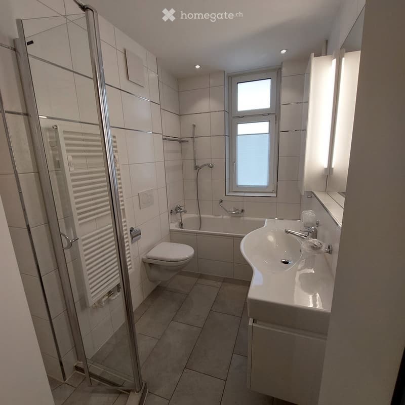 Modern bathroom with white tiles, a bathtub, toilet, sink, and a glass-enclosed shower area.