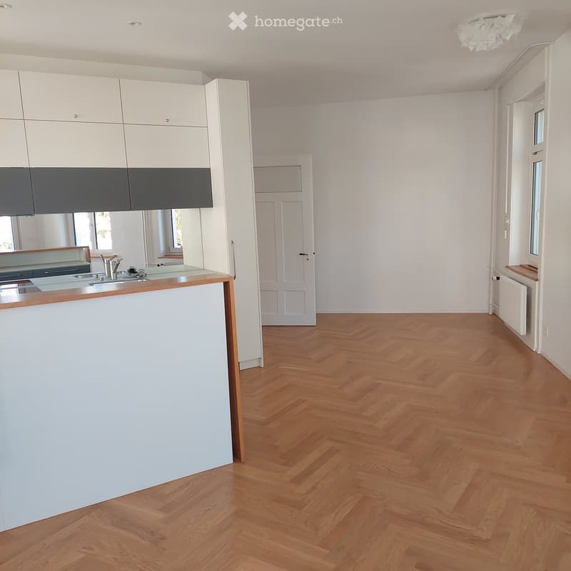Empty room with wooden herringbone flooring, white walls, and a white kitchen counter with sink and cabinets visible in the background.