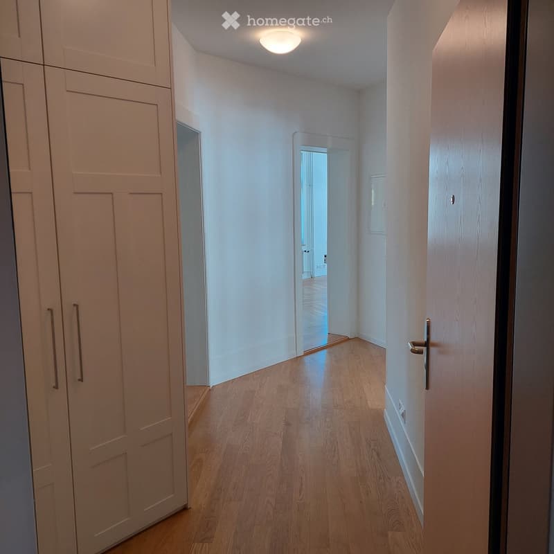 A narrow hallway with light wooden flooring, white walls, and a recessed ceiling light. Doors are visible on both sides, with one door slightly ajar, revealing a glimpse of another room.