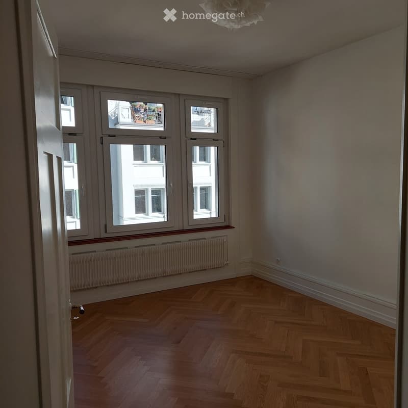 Empty room with wooden herringbone flooring, large windows, radiator, and white walls.