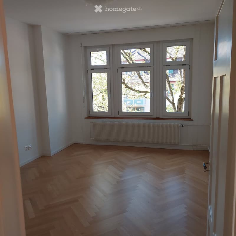 Empty room with wooden flooring, large windows, radiator, and visible electrical outlet.