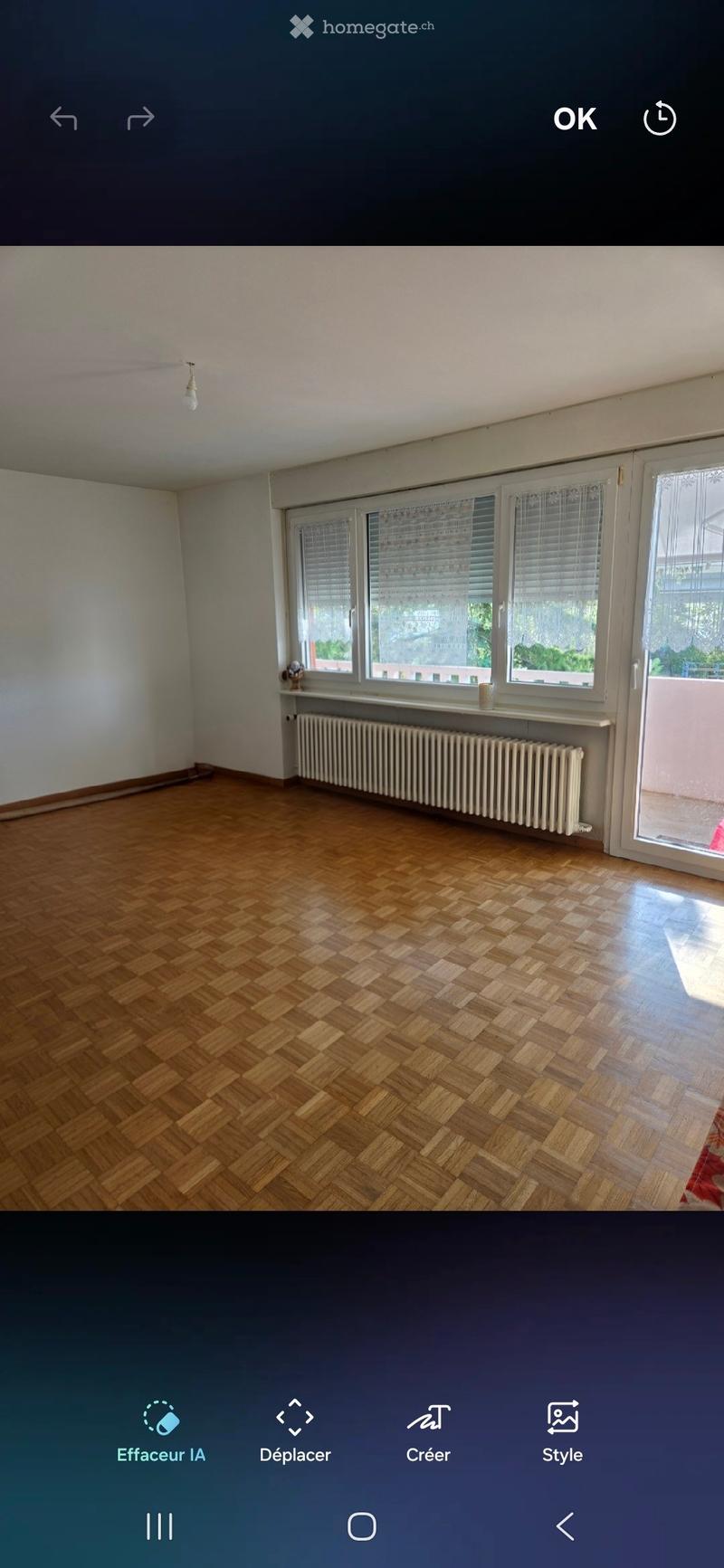 Empty room with wooden herringbone flooring, white walls, large windows with blinds, radiator, and a small shelf with a few decorative items.