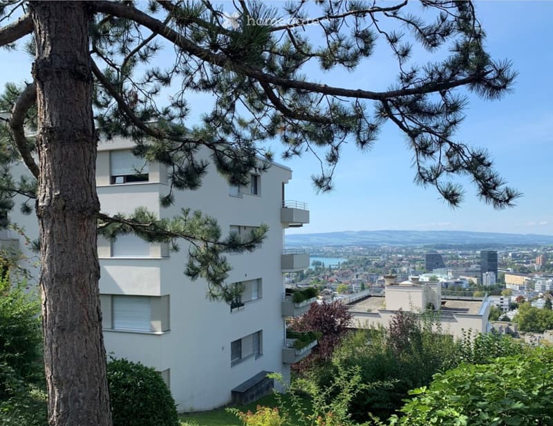 A modern white apartment building with multiple balconies, surrounded by trees and offering a panoramic city view.