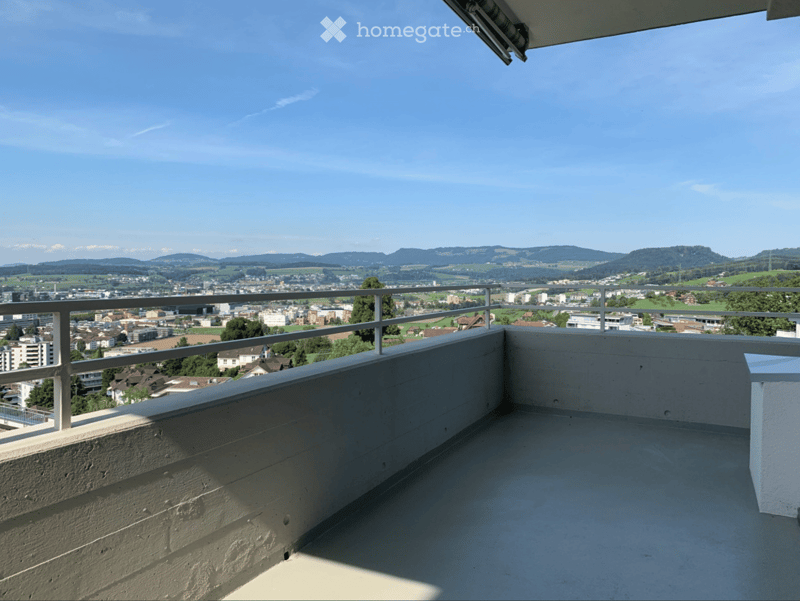Balcony with metal railing, overlooking a cityscape and distant mountains under a clear blue sky.