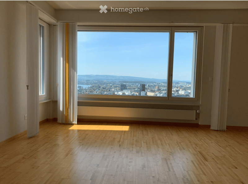 Empty room with wooden flooring, large windows offering a cityscape view, and light-colored walls.