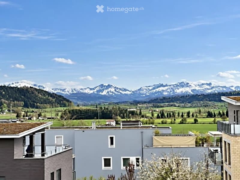 Modern residential building with balconies overlooking a scenic mountain landscape and green fields.