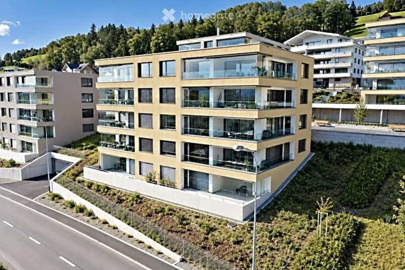 Modern multi-story residential building with balconies and large windows, surrounded by greenery and other buildings.