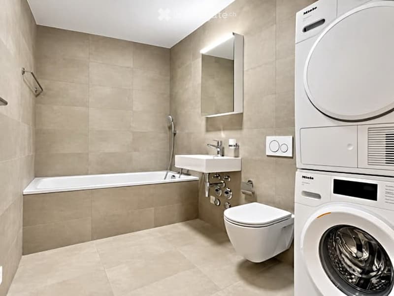Modern bathroom with beige tiles, bathtub, sink, toilet, mirror, and a private washing machine.