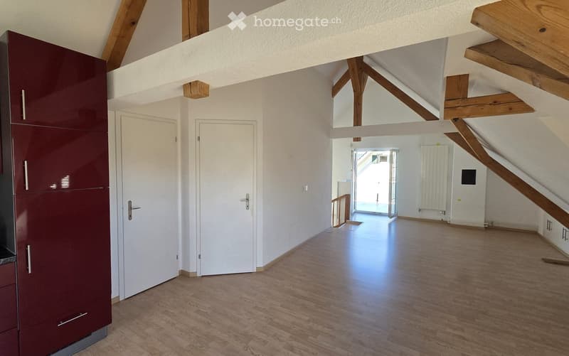 Empty room with wooden flooring, white walls, exposed wooden beams, and two white doors. The room is brightly lit and has a modern, minimalist design.