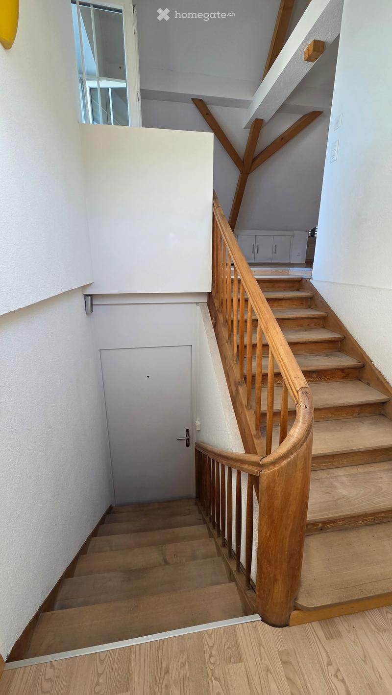 Wooden staircase with a wooden handrail and balusters, leading to an upper level. The walls are white, and there is a door at the end of the hallway.