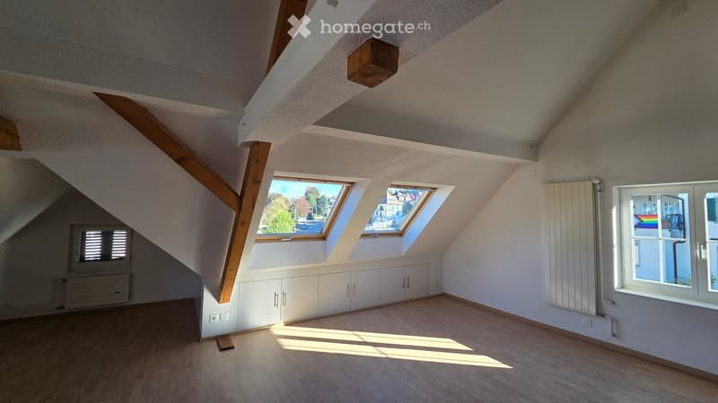 Bright, empty attic room with wooden beams, skylights, and white cabinetry along one wall.