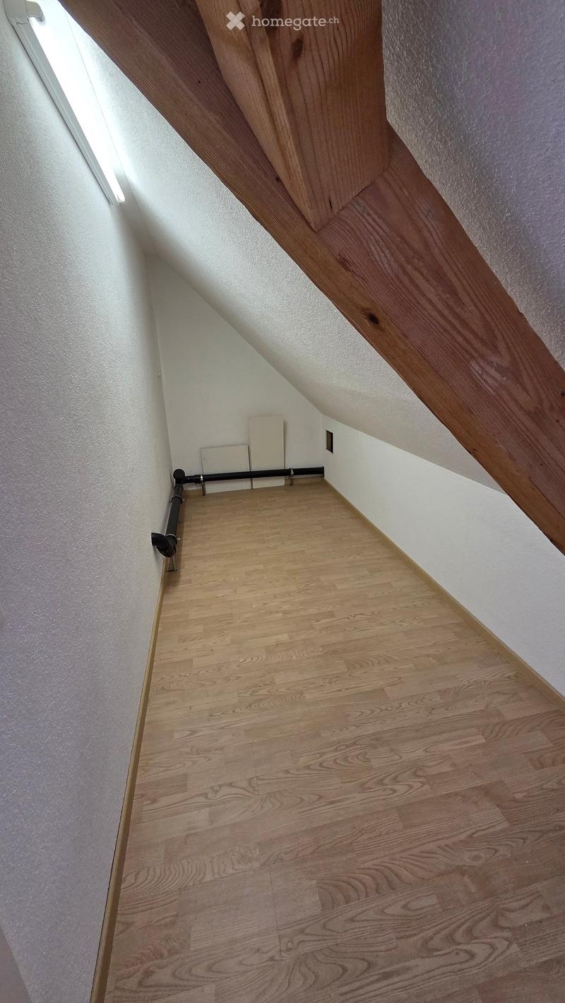 An attic space with wooden flooring, white walls, and exposed wooden beams. The room is empty with no furniture or equipment.