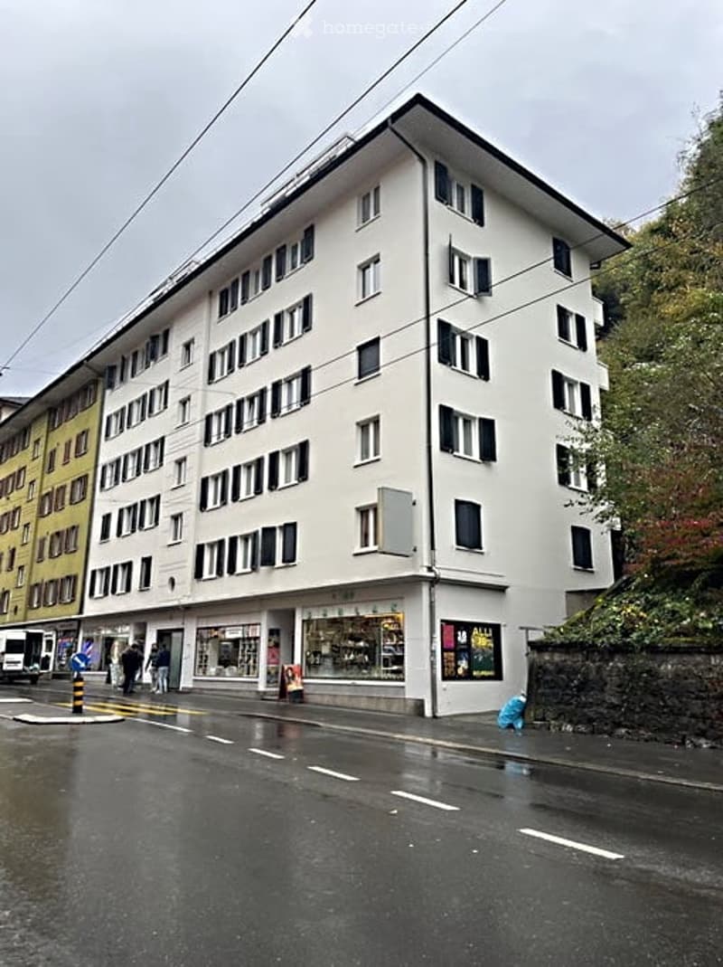 Multi-story white building with numerous windows and a storefront at the ground level, located on a wet street with pedestrians.