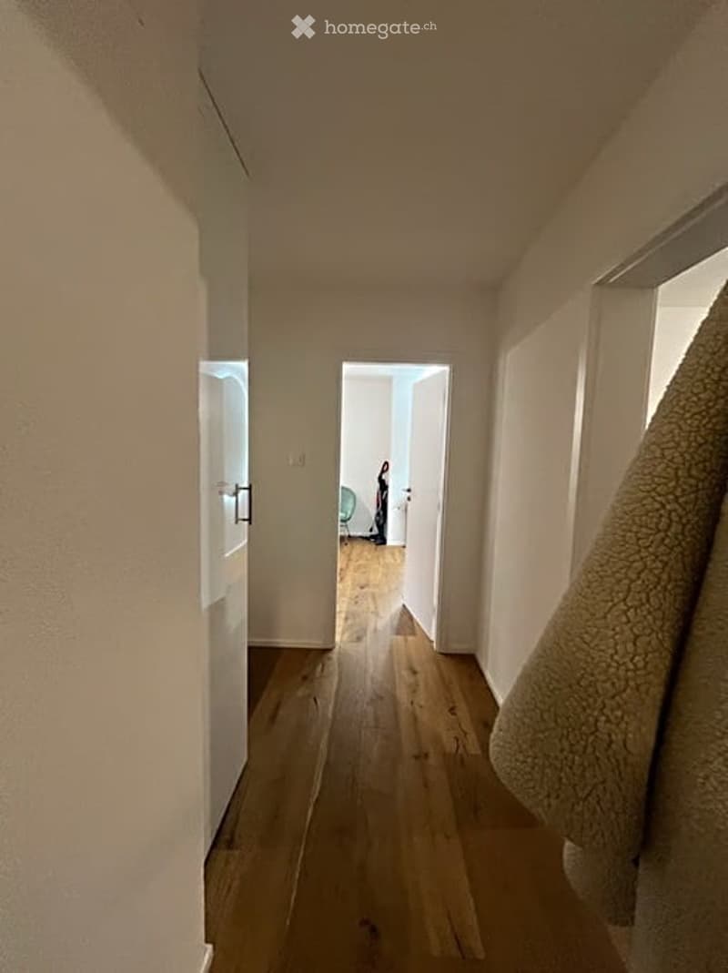 A narrow hallway with wooden flooring and white walls, leading to an open doorway that reveals a small room with a chair and a vacuum cleaner.