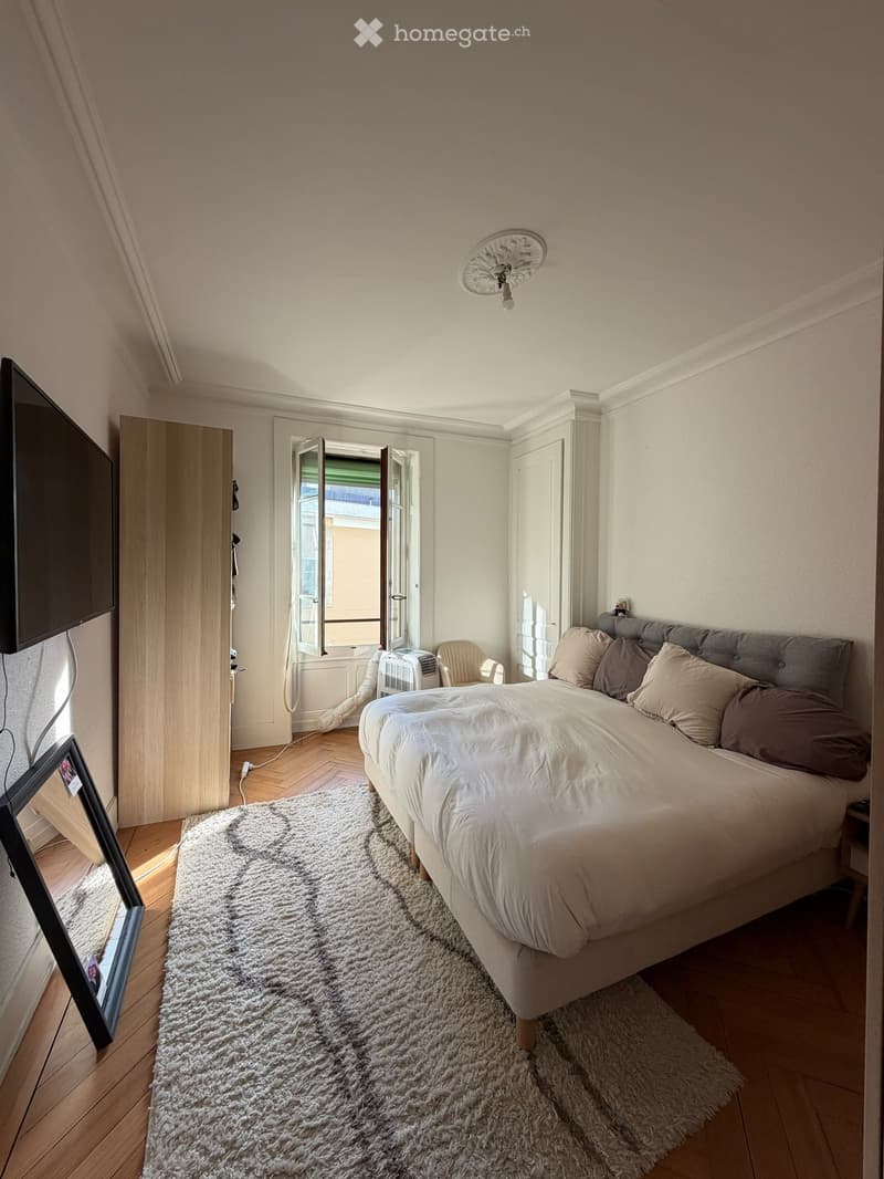 Cozy bedroom with a large bed, pillows, a fluffy rug, a chair, a screen, a wardrobe, and a fan. The room has wooden flooring and large windows with open shutters.