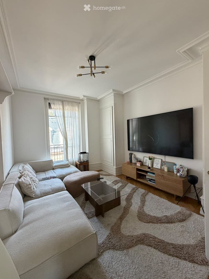 Modern living room with a grey sectional sofa, glass coffee table, ottoman, wooden side table, TV, and decorative items on a shelf.
