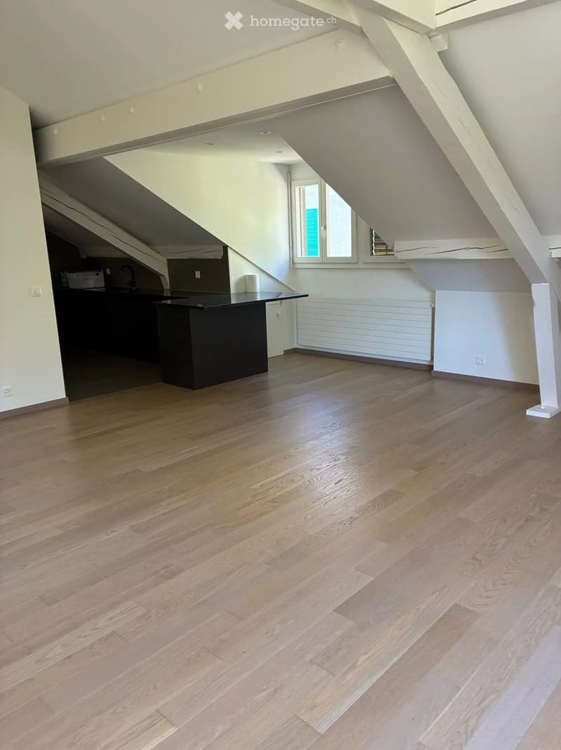 An empty attic space with wooden flooring, white walls, and exposed wooden beams. There is a black island in the background and a window letting in natural light.