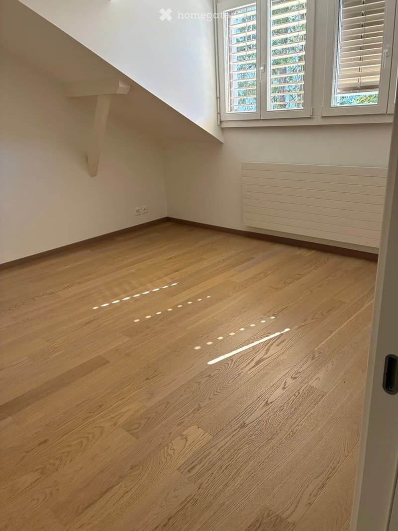 Empty room with wooden flooring, white walls, and a window with blinds. The floor features integrated LED lighting strips along the edges.