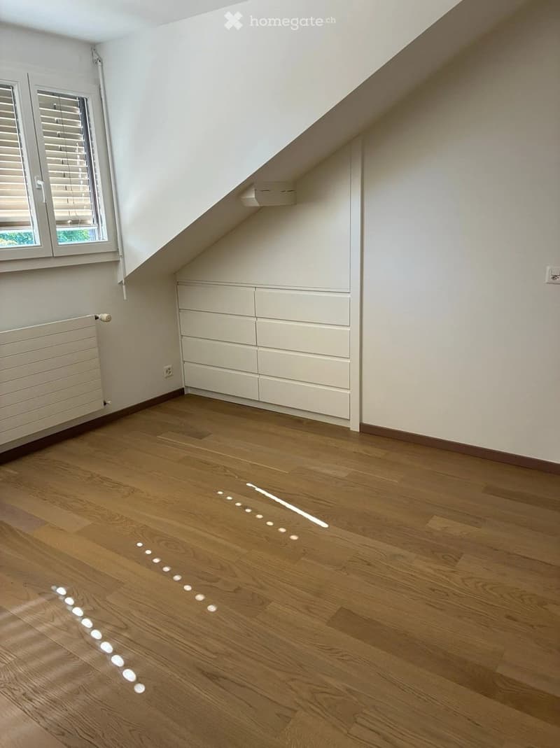 Empty attic room with wooden flooring, white walls, built-in storage cabinets, and a window with blinds.
