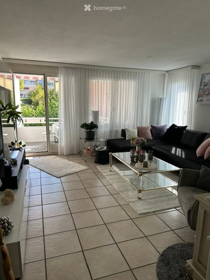 Modern living room with large windows and balcony access, featuring a black sectional sofa, glass coffee table, side table with plant, and television on a stand.