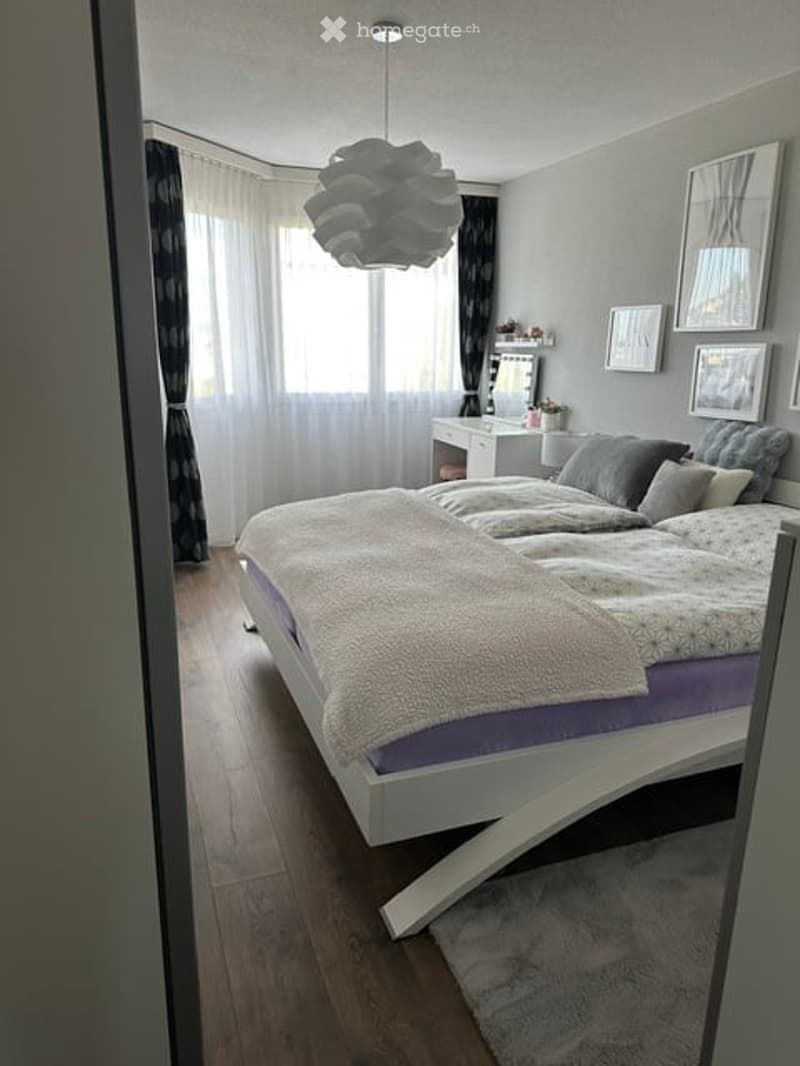 Modern bedroom with a white platform bed, grey bedding, a vanity with a mirror, and picture frames on the wall. The room features wooden flooring and large windows with white curtains.