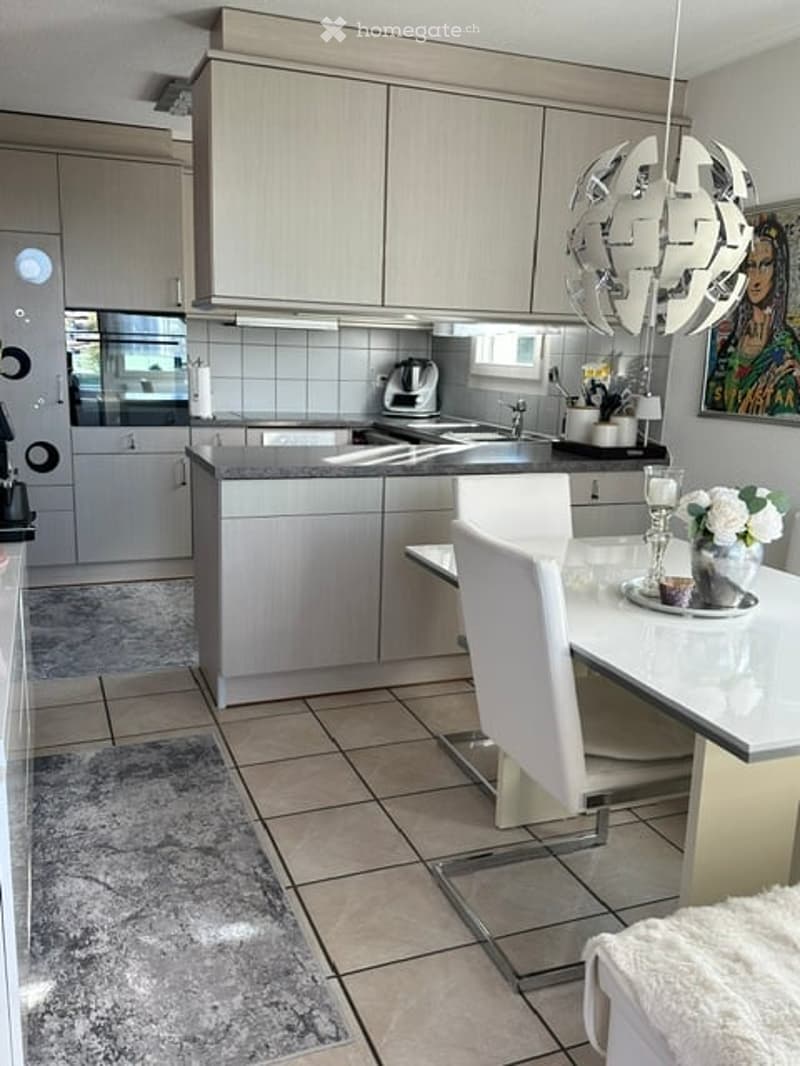 Modern kitchen with light grey cabinets, a central island, a sink, a coffee maker, and a dining area with a white table and chairs.