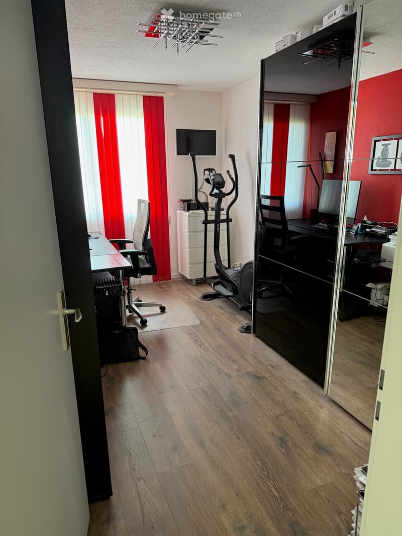 A modern, well-lit room with wooden flooring, featuring a desk with a chair, a fitness bike, a screen, and a large mirror. The room has red and white curtains and a white cabinet.