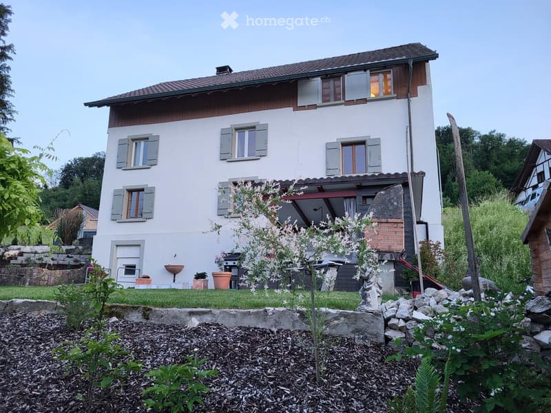 Two-story house with white walls, wooden shutters, and a tiled roof. The house features a well-maintained garden with various plants, a small patio area, and a barbecue setup.