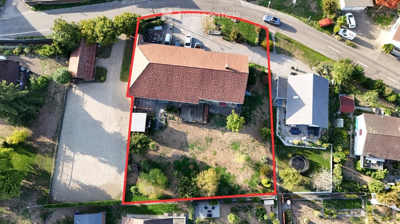Aerial view of a two-story house with a tiled roof, surrounded by a garden and driveway with multiple parked cars.