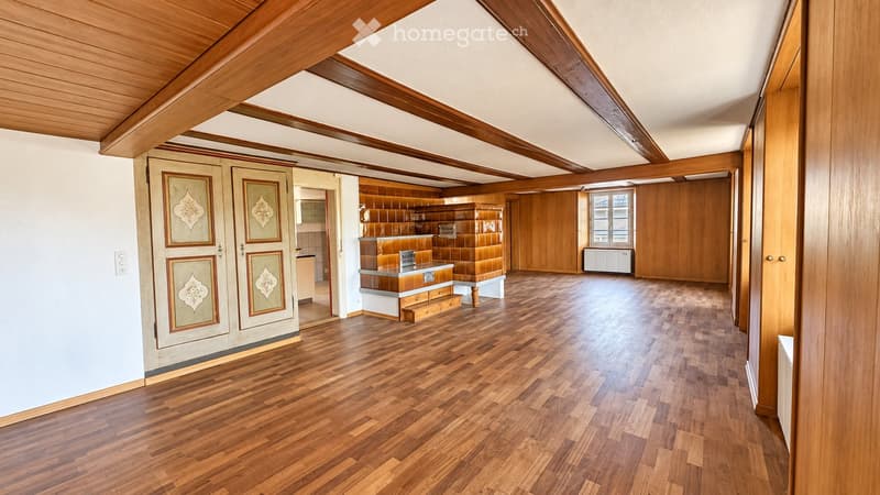 Spacious, unfurnished room with wooden flooring, exposed wooden beams on the ceiling, and a small kitchen area visible in the background.