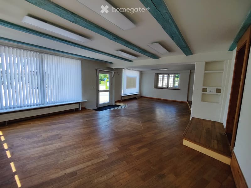Empty room with wooden flooring, white walls, and large windows with blinds. The space features exposed wooden beams on the ceiling and a door leading to an outdoor area.