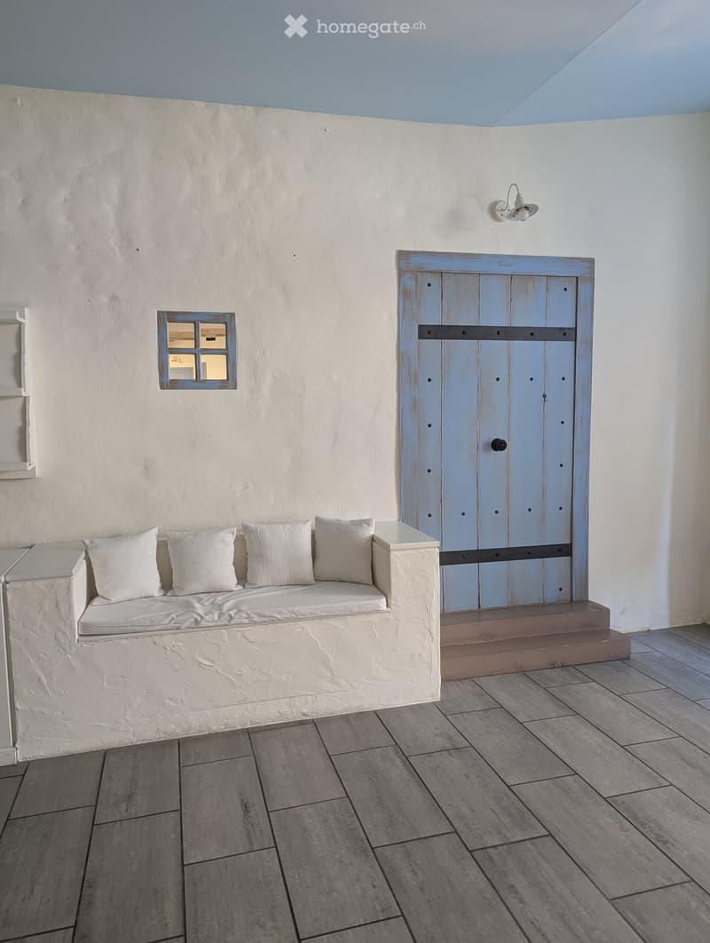 The image shows an empty bench with three pillows, white walls, a window on the left, and a wooden door on the right with a small window at the top.
