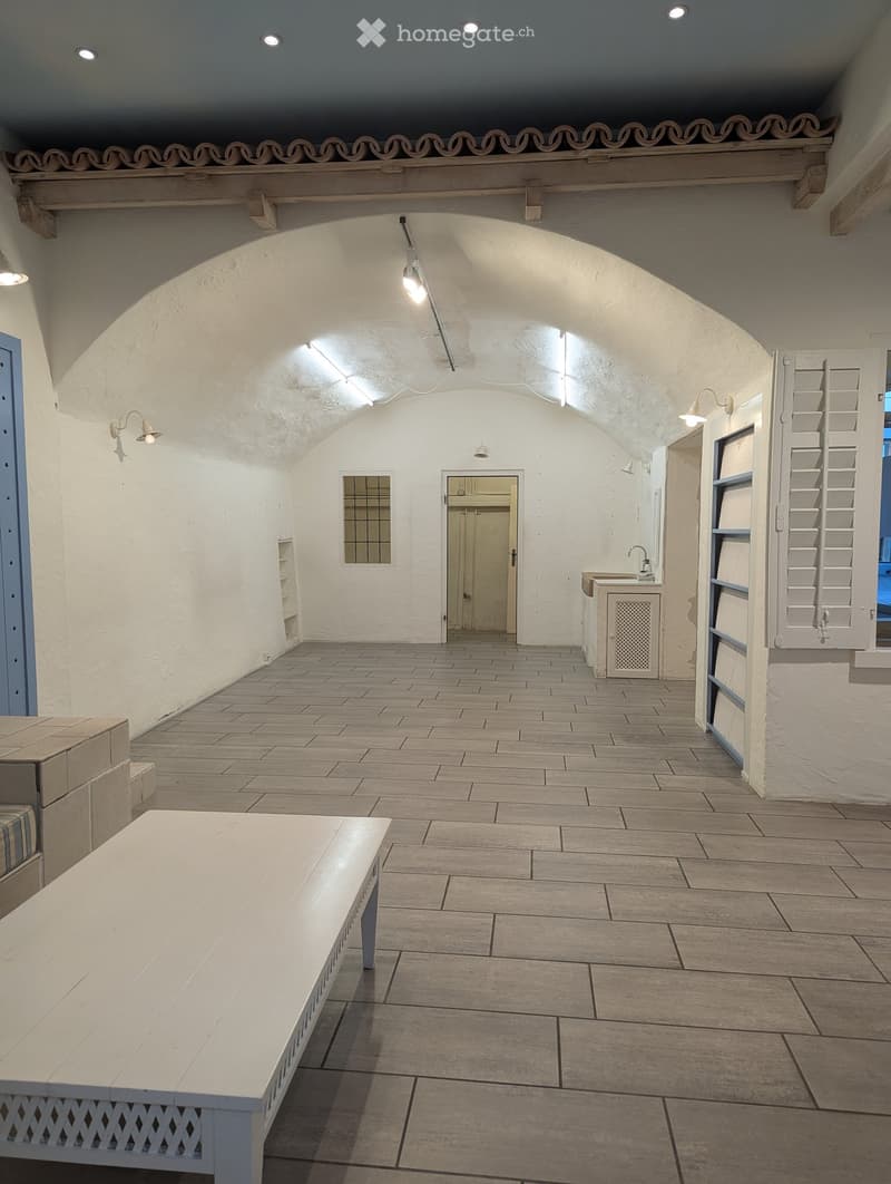 A spacious room with an arch ceiling and gray tile floor. On the left is a door with shelves above it and wall-mounted lights. In the middle is a doorway with an open door to a sink area. On the right, there are white shutters and another doorway. A white