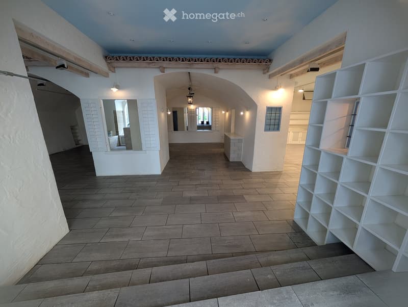 Spacious, well-lit hallway with tiled flooring, white walls, and wooden beams. Features an arched doorway leading to another room, built-in shelving unit on the right, and a small bench near the doorway.