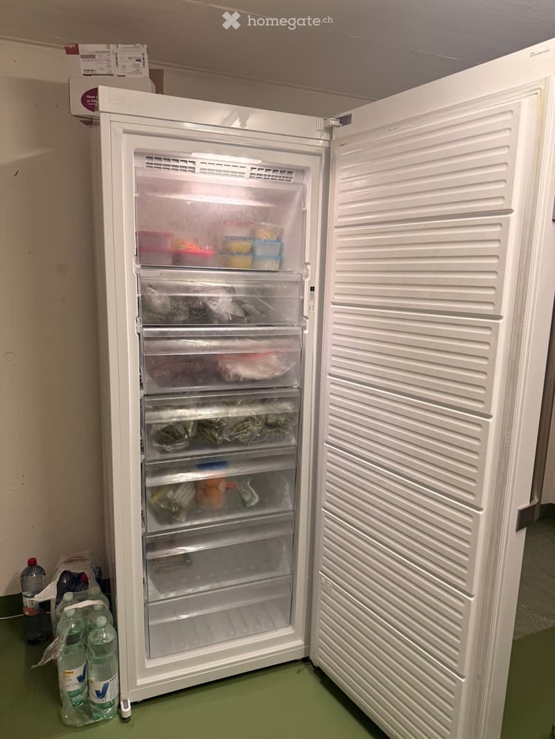 A white refrigerator with multiple shelves and drawers is open, showing various food items inside. Several water bottles are placed on the floor next to the refrigerator.