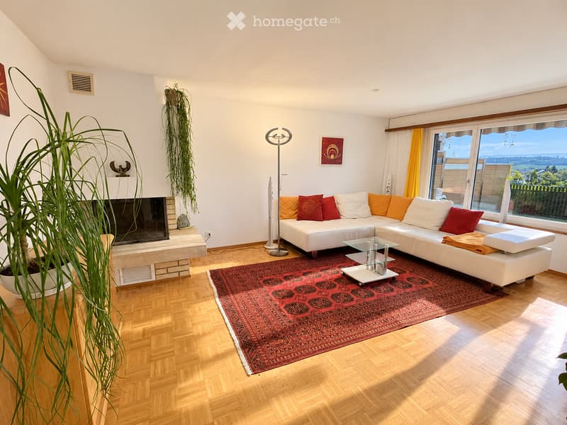 Modern living room with white sectional sofa, red and orange cushions, glass coffee table, large television, floor lamp, and a view through large windows.