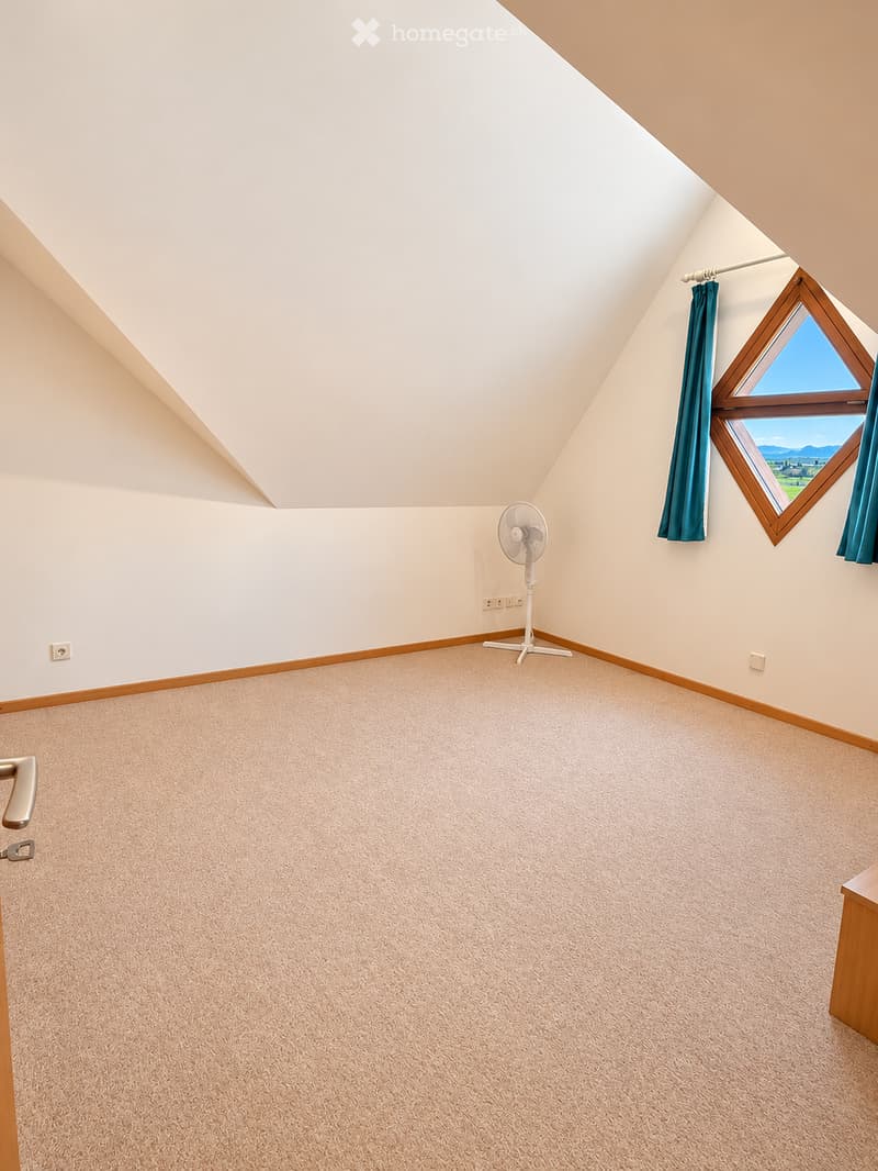 Empty attic room with sloped ceilings, carpeted floor, white walls, and a single window with blue curtains. A small white fan is placed near the wall.
