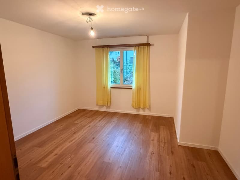 Empty room with wooden flooring, white walls, and a window with yellow curtains.