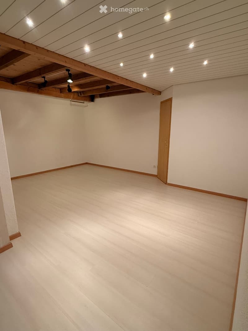 Empty room with wooden floor, white walls, exposed wooden beams on the ceiling, and a single wooden door.