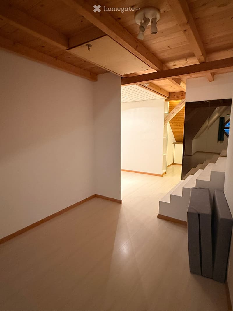 Empty hallway with light-colored wooden floor, white walls, and exposed wooden ceiling beams. A staircase leads upwards, and a mirror is mounted on the wall.