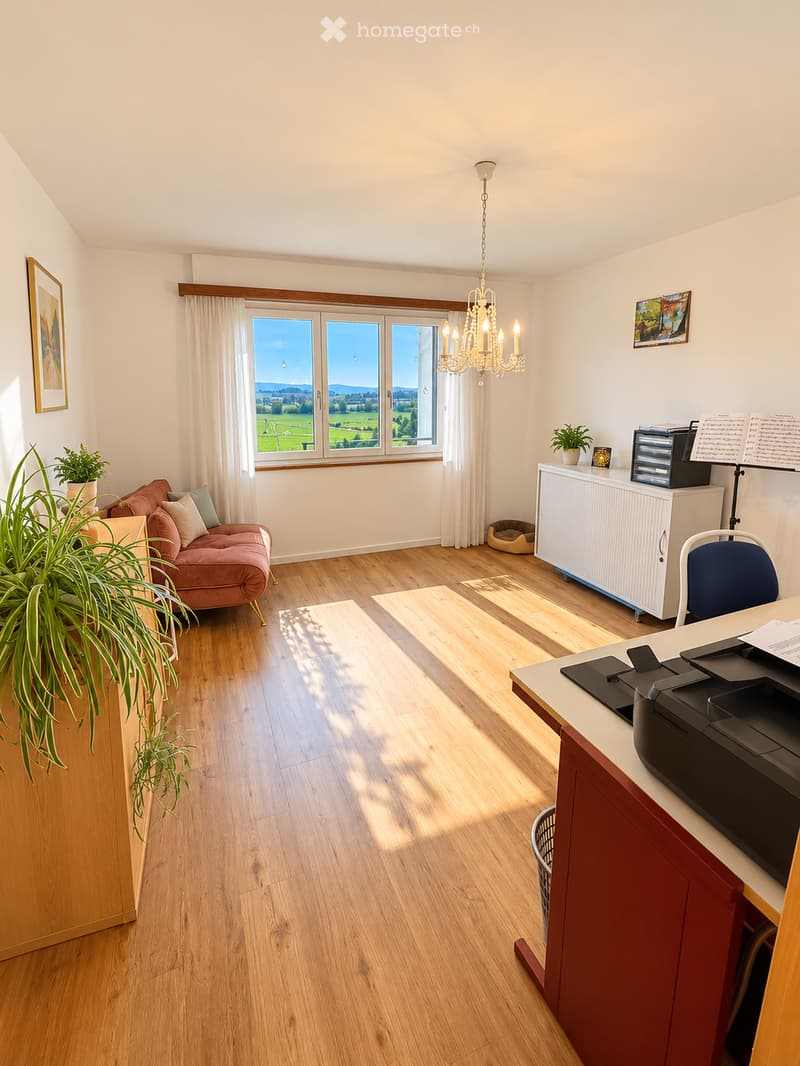 Bright room with wooden flooring, large windows overlooking a scenic landscape, a pink sofa, a desk with a printer, a chandelier, and decorative plants.