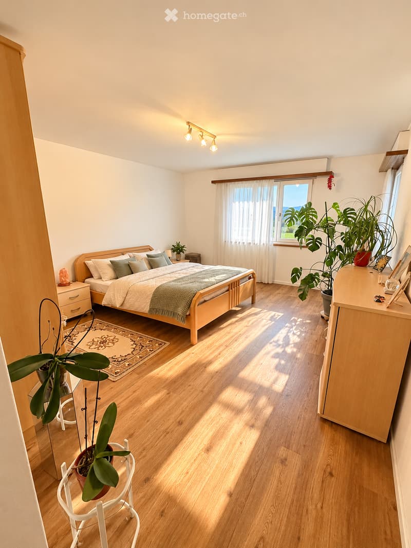 Bright bedroom with wooden flooring, large window with white curtains, bed with pillows and blanket, nightstand, chair, and potted plants.