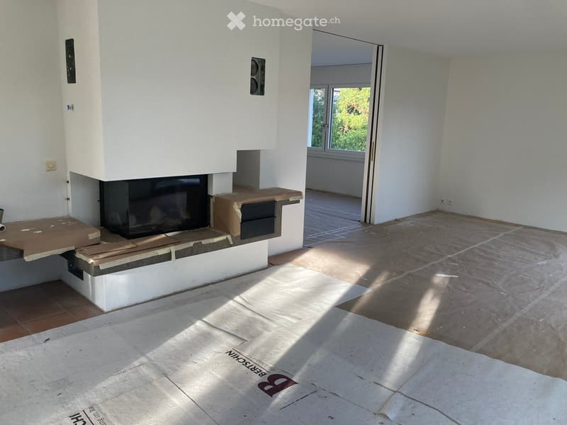 Empty room with white walls, tiled floor, and a fireplace area with protective coverings. Sunlight streams in from a window on the right side.