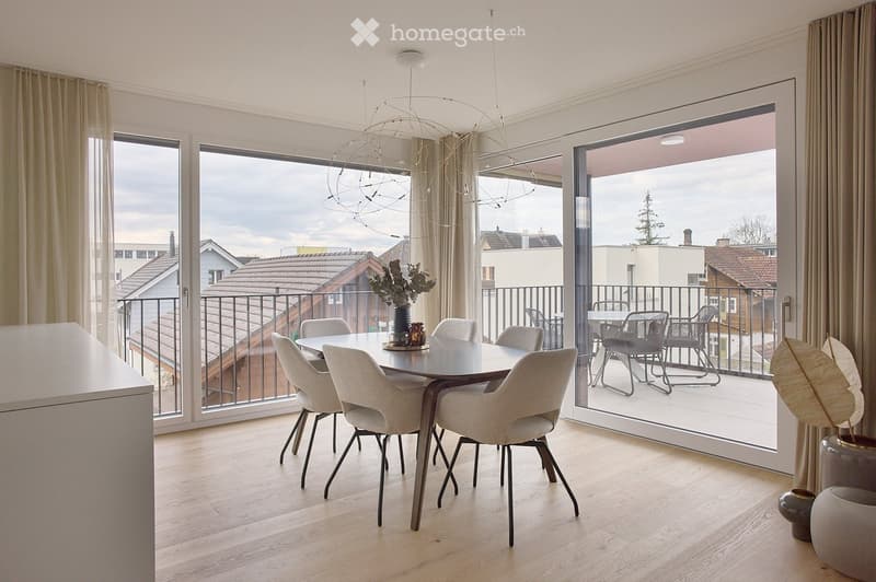 Modern dining room with light wood flooring, large windows, and a round dining table with six chairs. A decorative vase with greenery sits on the table, and a balcony with outdoor furniture is visible through the sliding glass doors.