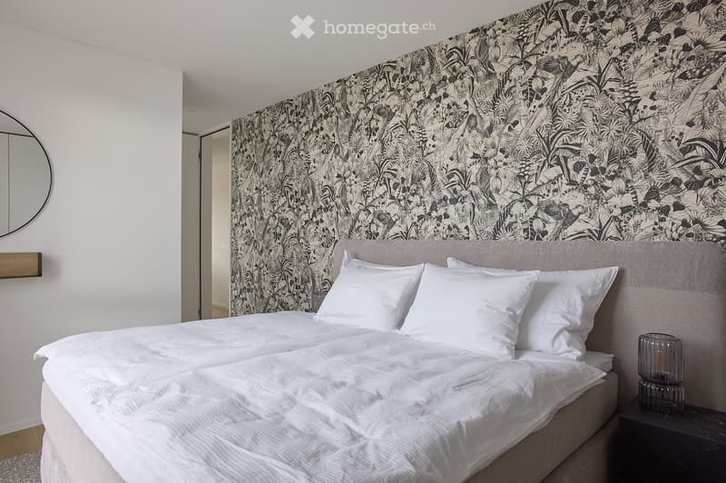 Modern bedroom with a large bed, white bedding, a decorative headboard, and a patterned accent wall. A nightstand with a table lamp is visible, along with a round mirror on the wall.