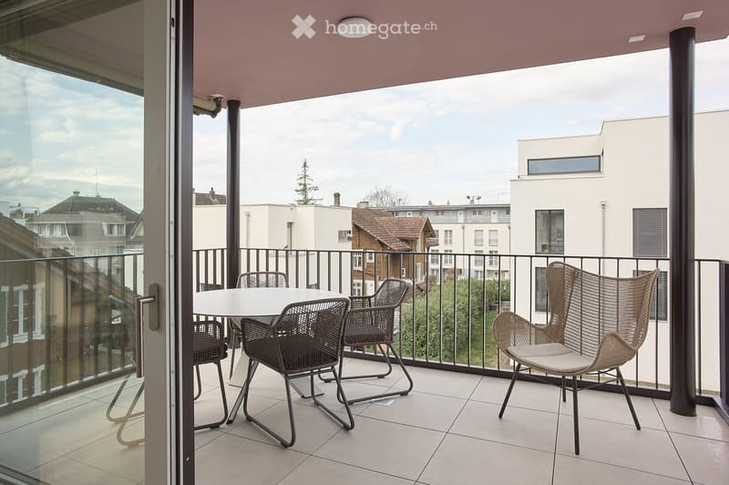 Balcony with a white table and four wicker chairs, overlooking a cityscape with buildings and trees.