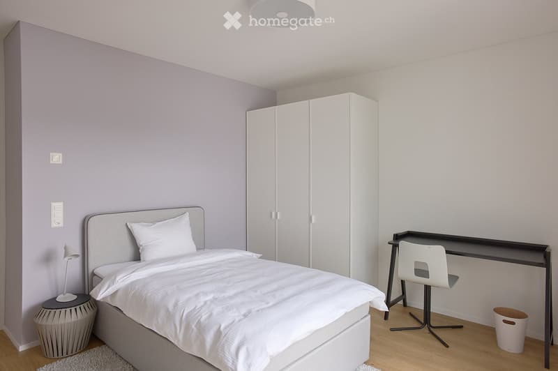 Modern, minimalist bedroom with a white bed, desk, chair, wardrobe, and a small lamp on a stool.