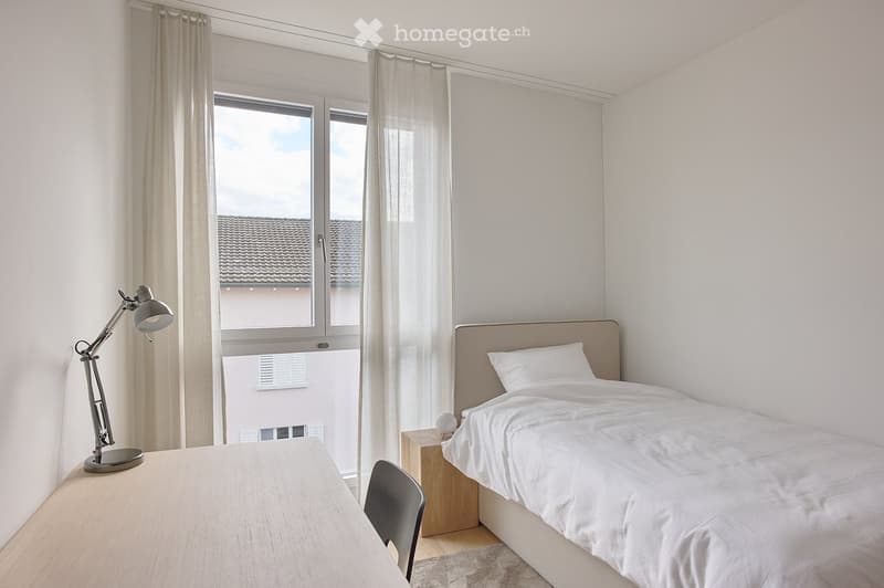 A modern, minimalist bedroom with a large bed, nightstand, desk, and chair. The room features large windows with sheer curtains, allowing natural light to fill the space.