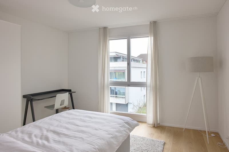 Modern, minimalist bedroom with a white bed, a sleek black desk, a white chair, and a tall floor lamp near the window.