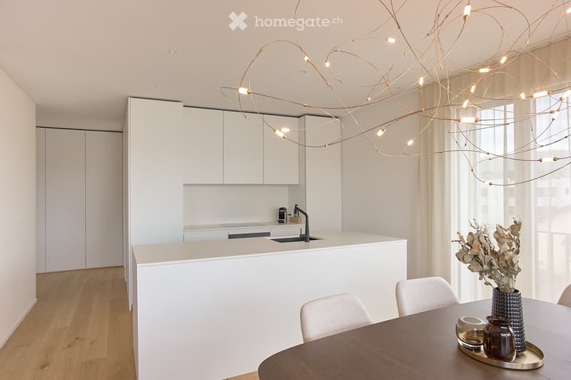 Modern kitchen and dining area with white cabinets, a central island, a round wooden dining table with chairs, and a decorative vase on the table. The space features a unique hanging light fixture and large windows with sheer curtains.
